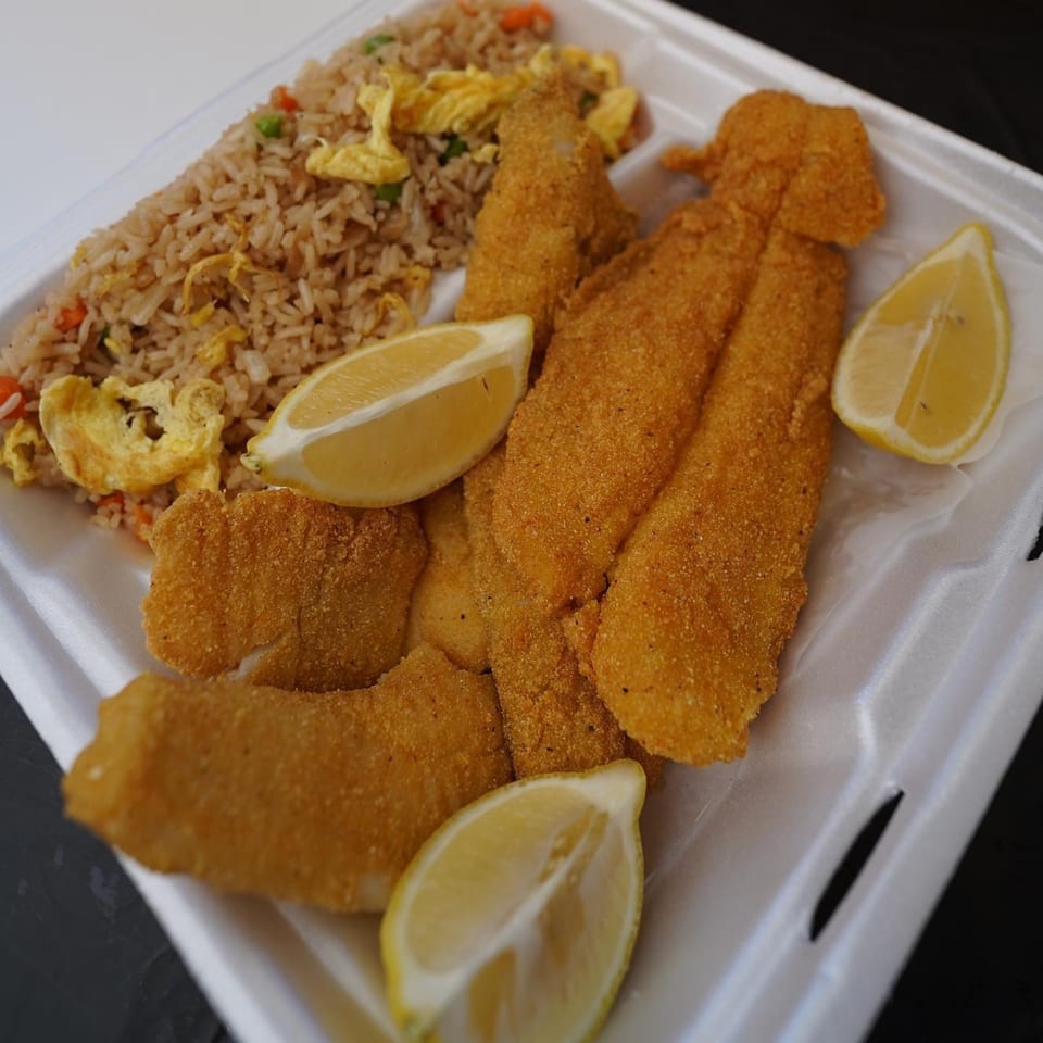 Fried fish fillets with lemon wedges and fried rice in a white takeout container.